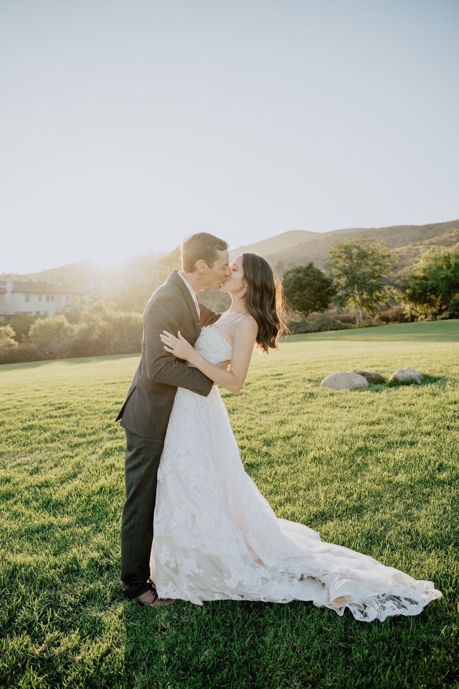 A couple in wedding attire share romantic moments in a sunlit green field at sunset with mountains in the background.