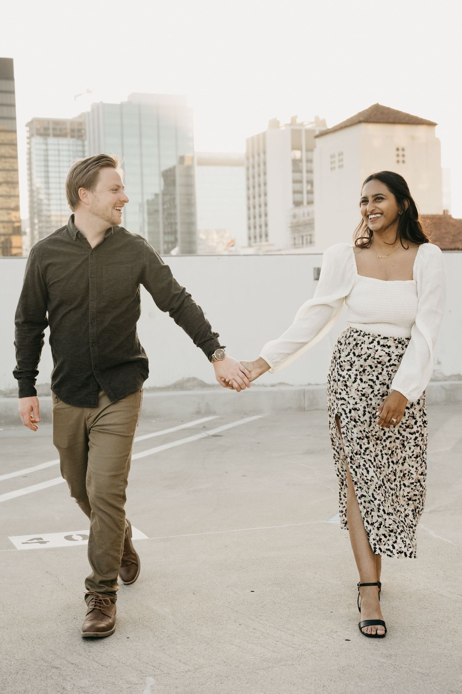 A couple holds hands and dances playfully on a rooftop with city buildings visible in the background.
