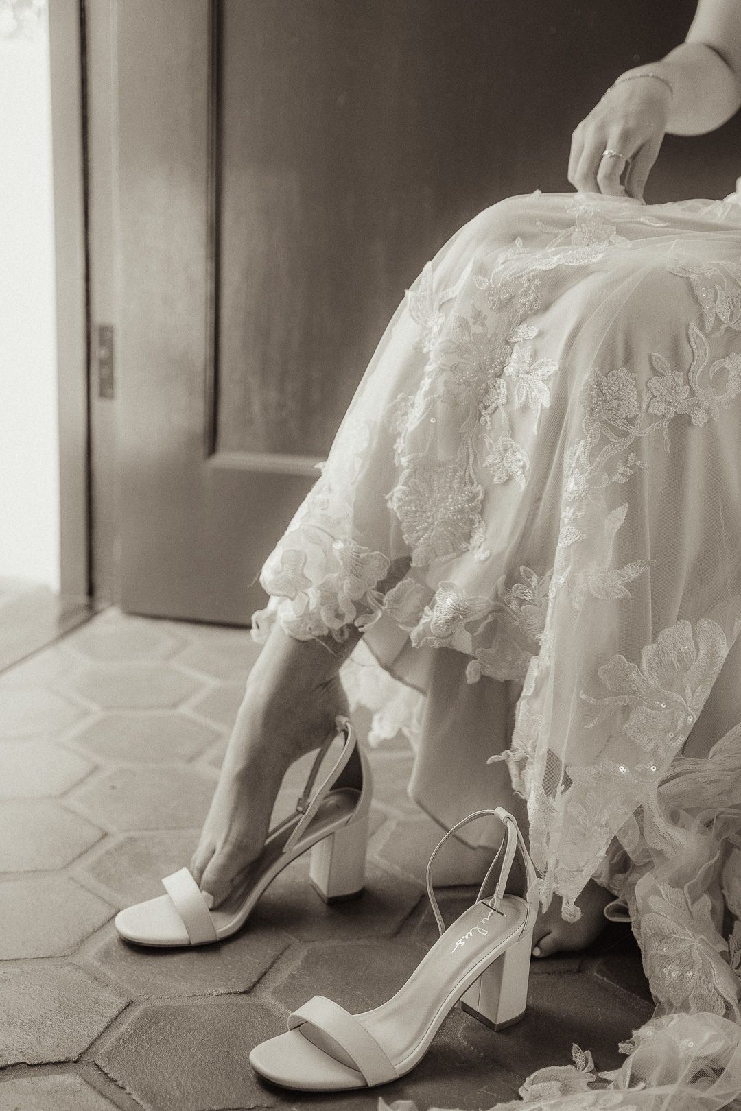 A close-up black and white photo of white bridal shoes and lace wedding dress hem on hexagonal tile floor.