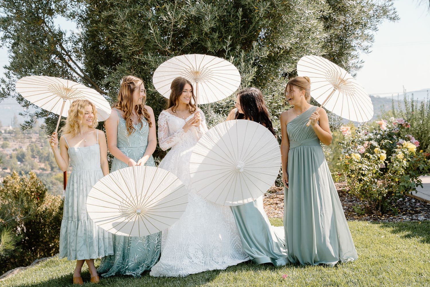 Wedding party members in light blue dresses twirl white parasols on a sunny hillside during an outdoor celebration.