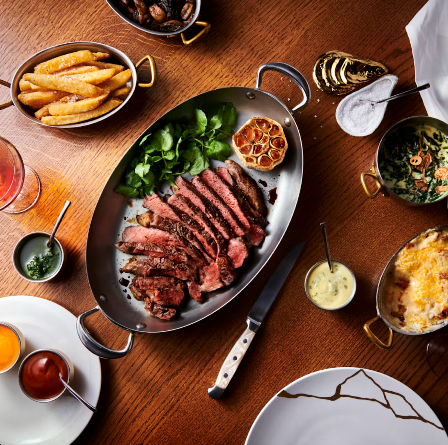 Juicy sliced steak served on dark plates with sides of french fries and fresh vegetables on a wooden table.