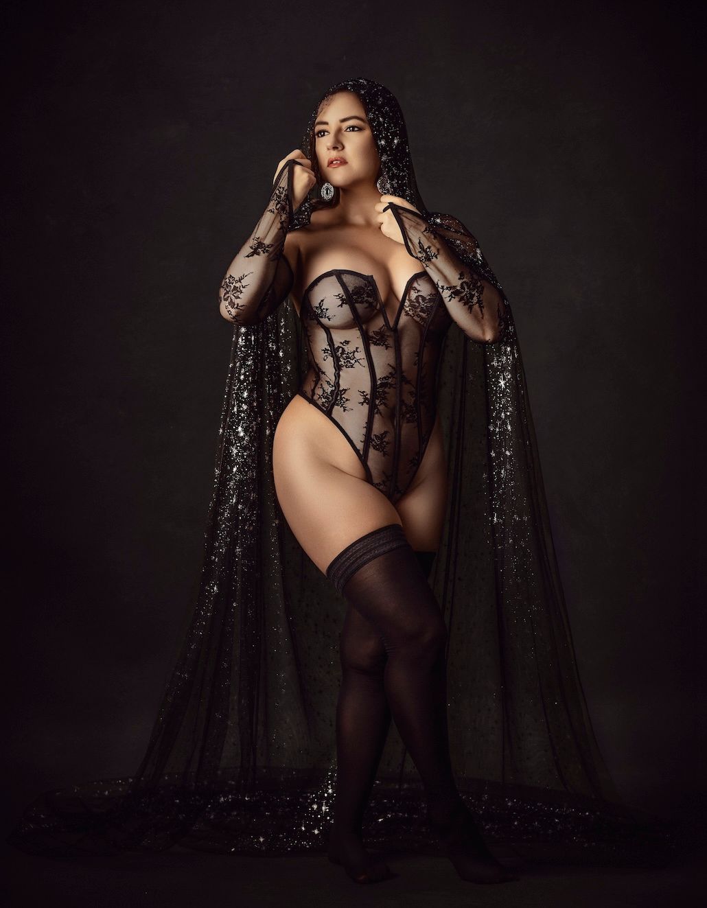 Woman in black lace bodysuit and sequined cape poses elegantly against dark studio background.