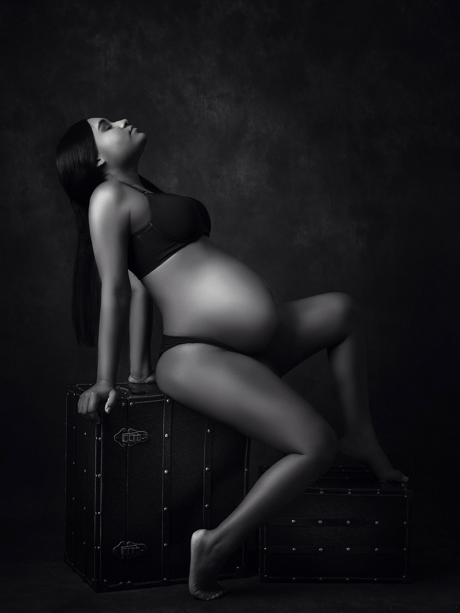 Dramatic monochrome maternity portrait with figure reclining on vintage luggage.