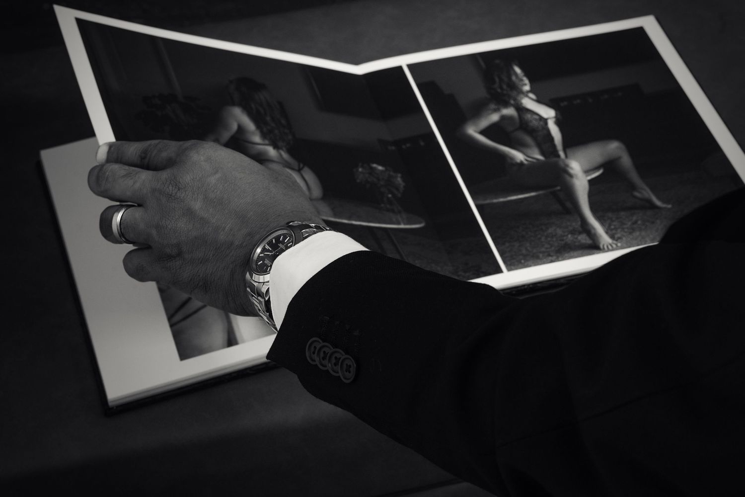 Man in suit wearing luxury watch examines open photography book with black and white artistic images.
