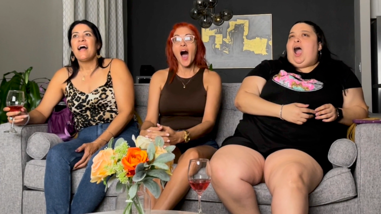 Group of friends laughing and socializing on grey couch with flowers and drinks.