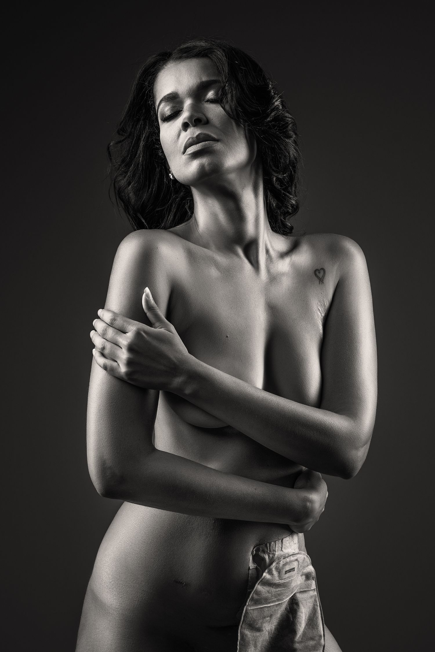 Artistic black and white portrait of a woman with dark hair in an intimate pose against dark background. Crohn's disease Boudoir Photo