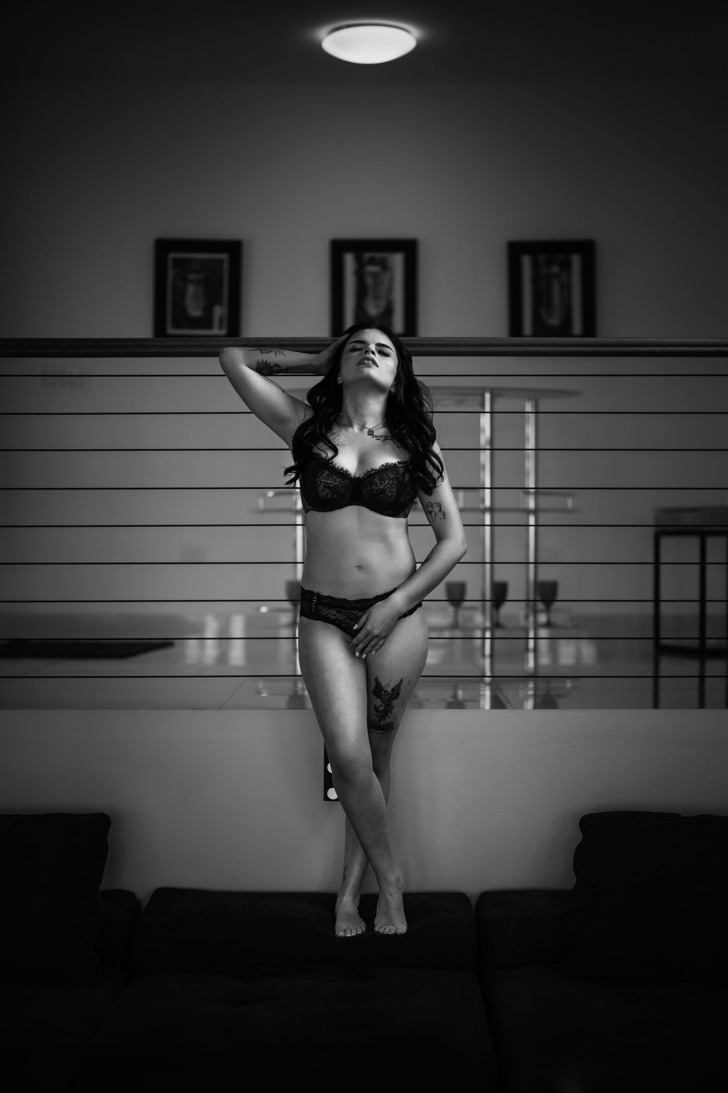 Black and white boudoir photo of a woman in lingerie posing confidently in a dimly lit modern interior.