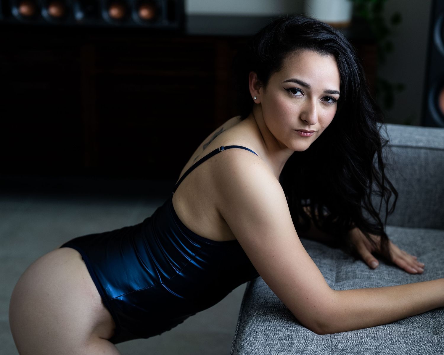 Woman in blue satin bodysuit posing on gray sofa in moody indoor lighting.