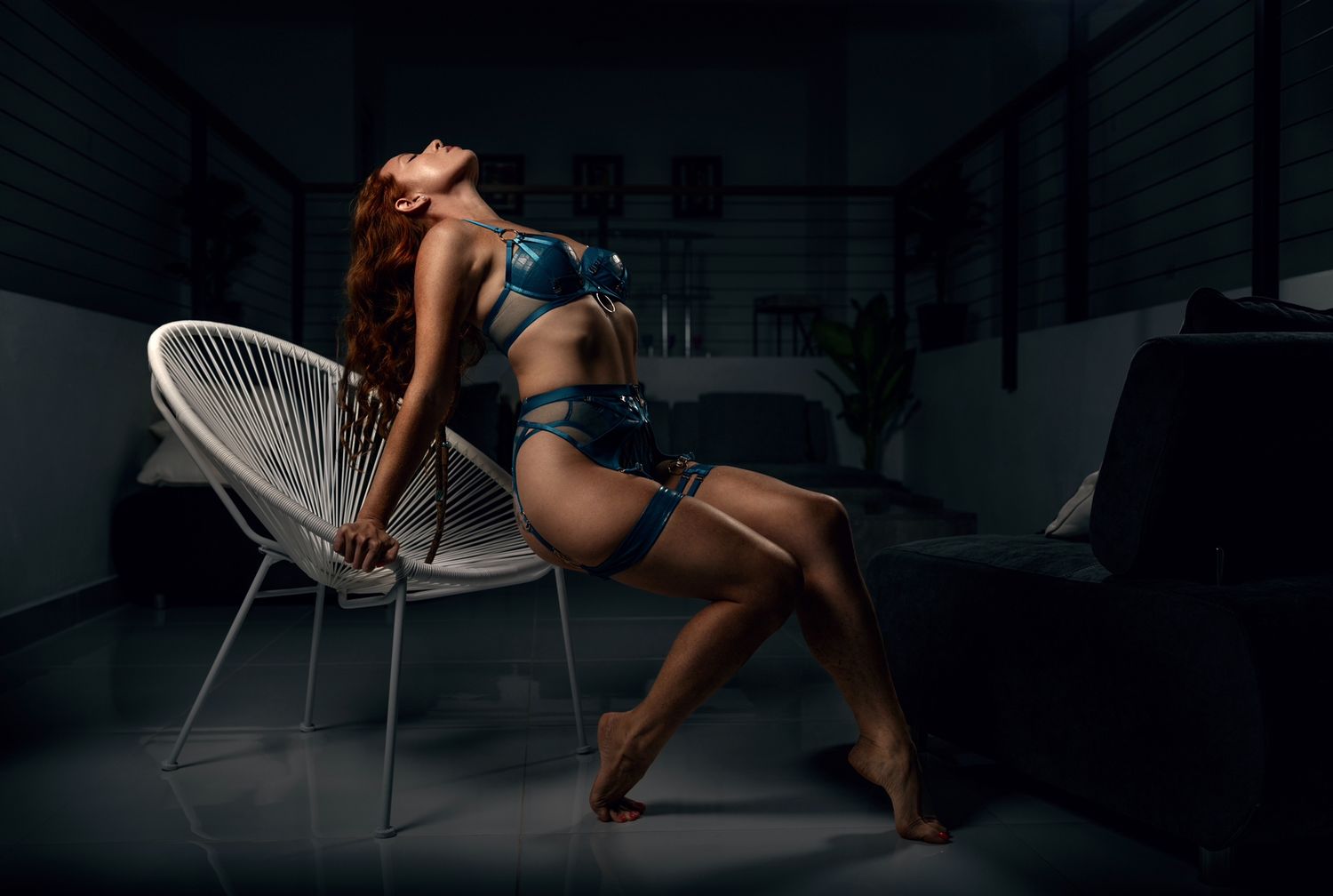Woman in blue lingerie posing gracefully beside a white chair in a dimly lit modern room.