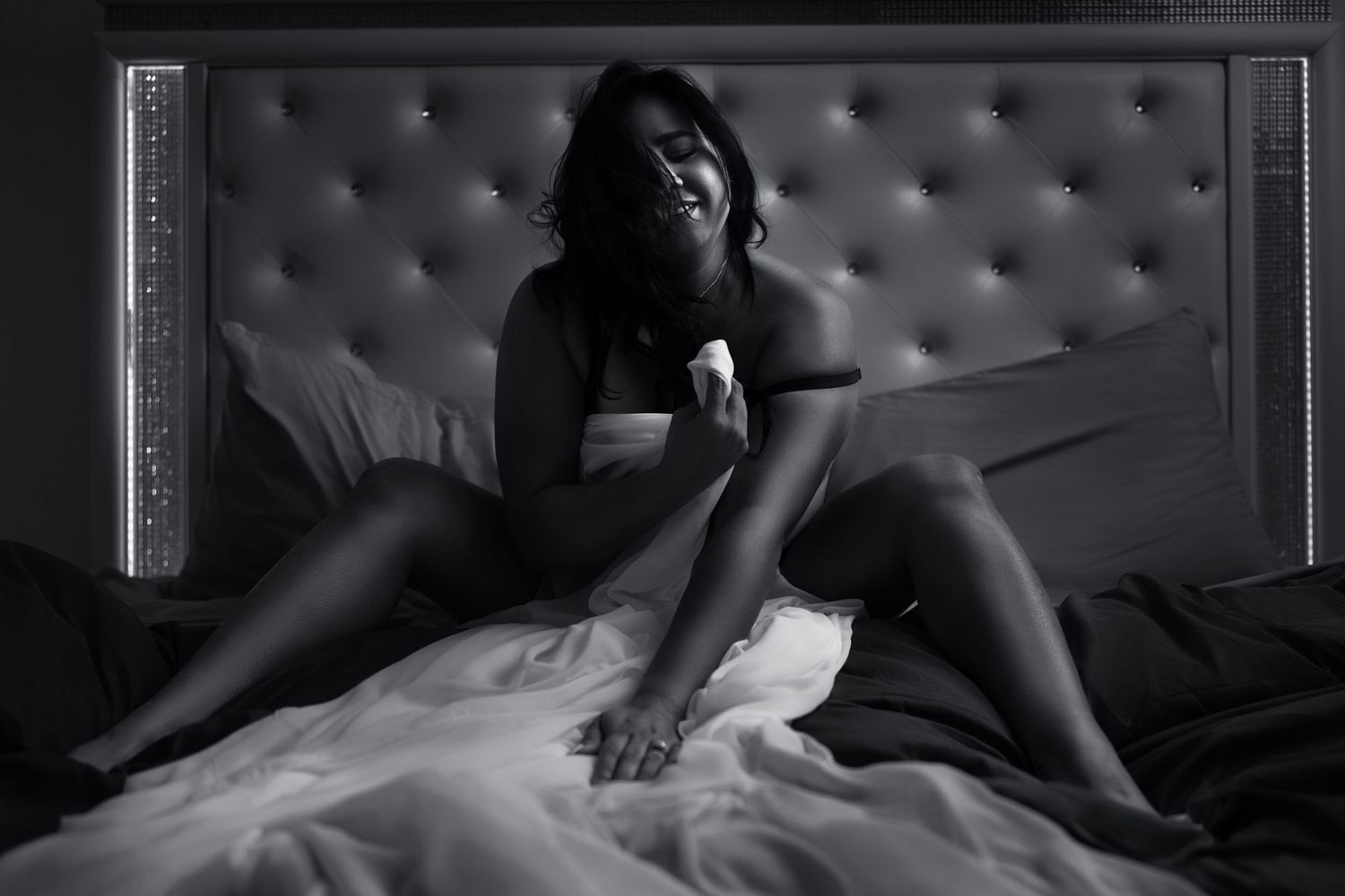 Black and white boudoir photo of a woman sitting on a bed with white sheets and tufted headboard.