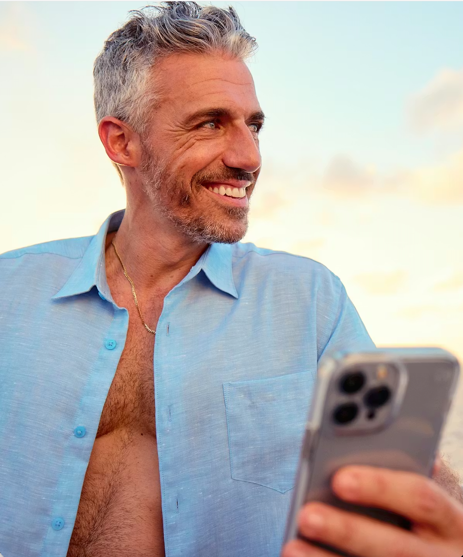 A person with gray hair wearing a blue shirt smiles while looking at their smartphone outdoors during sunset.