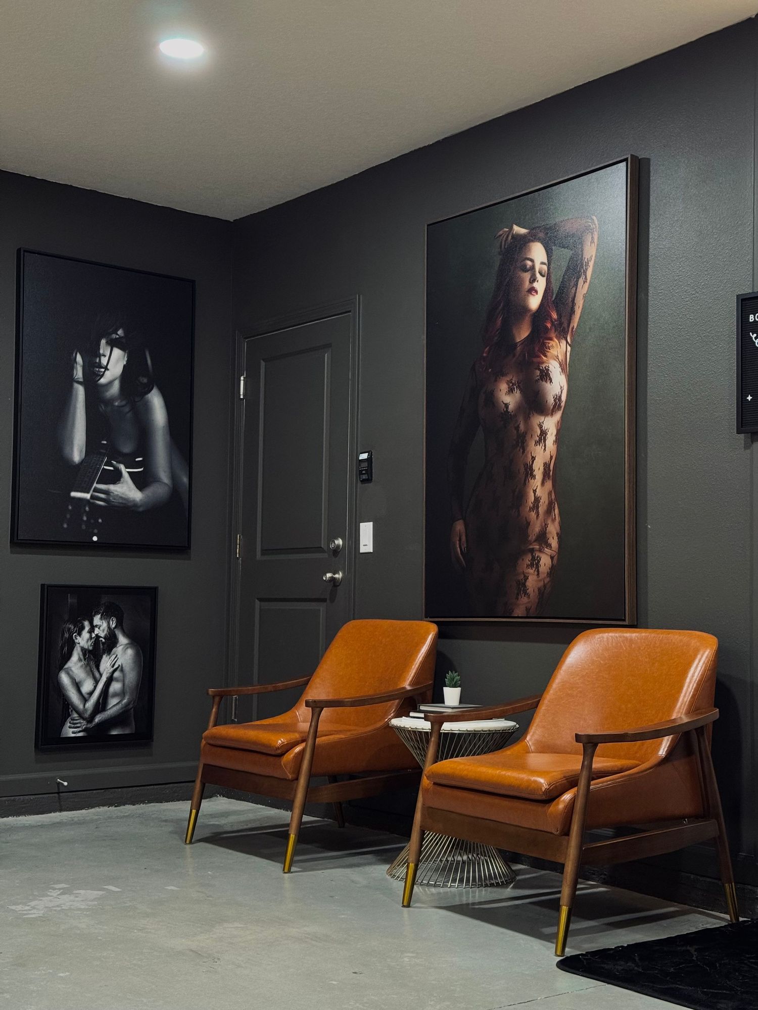 Two tan leather mid-century modern chairs flank a small table in a dark gray room with large framed fine art photography on walls.