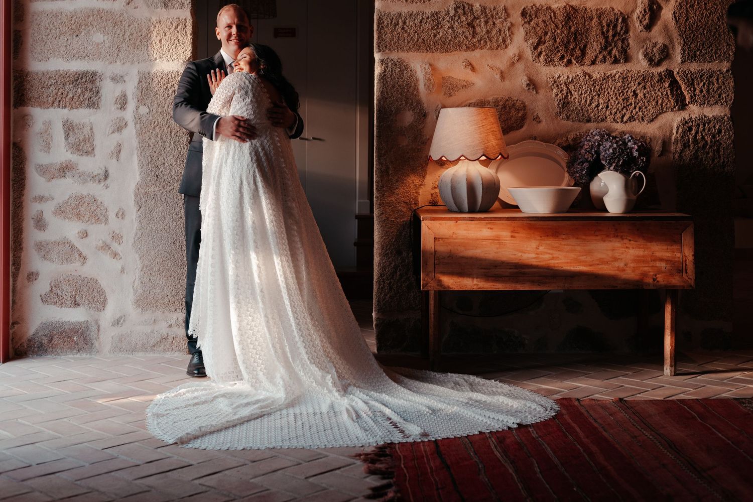 Bride and groom share an intimate moment in a rustic stone room, her long white gown trailing on a red rug.