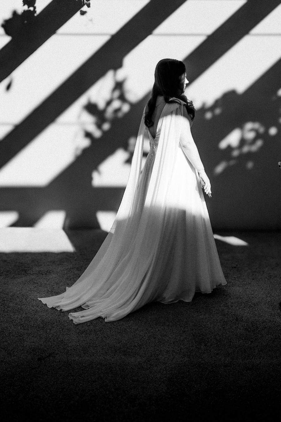 Black and white photo of a woman in a flowing white gown standing against a wall with dramatic diagonal shadows.