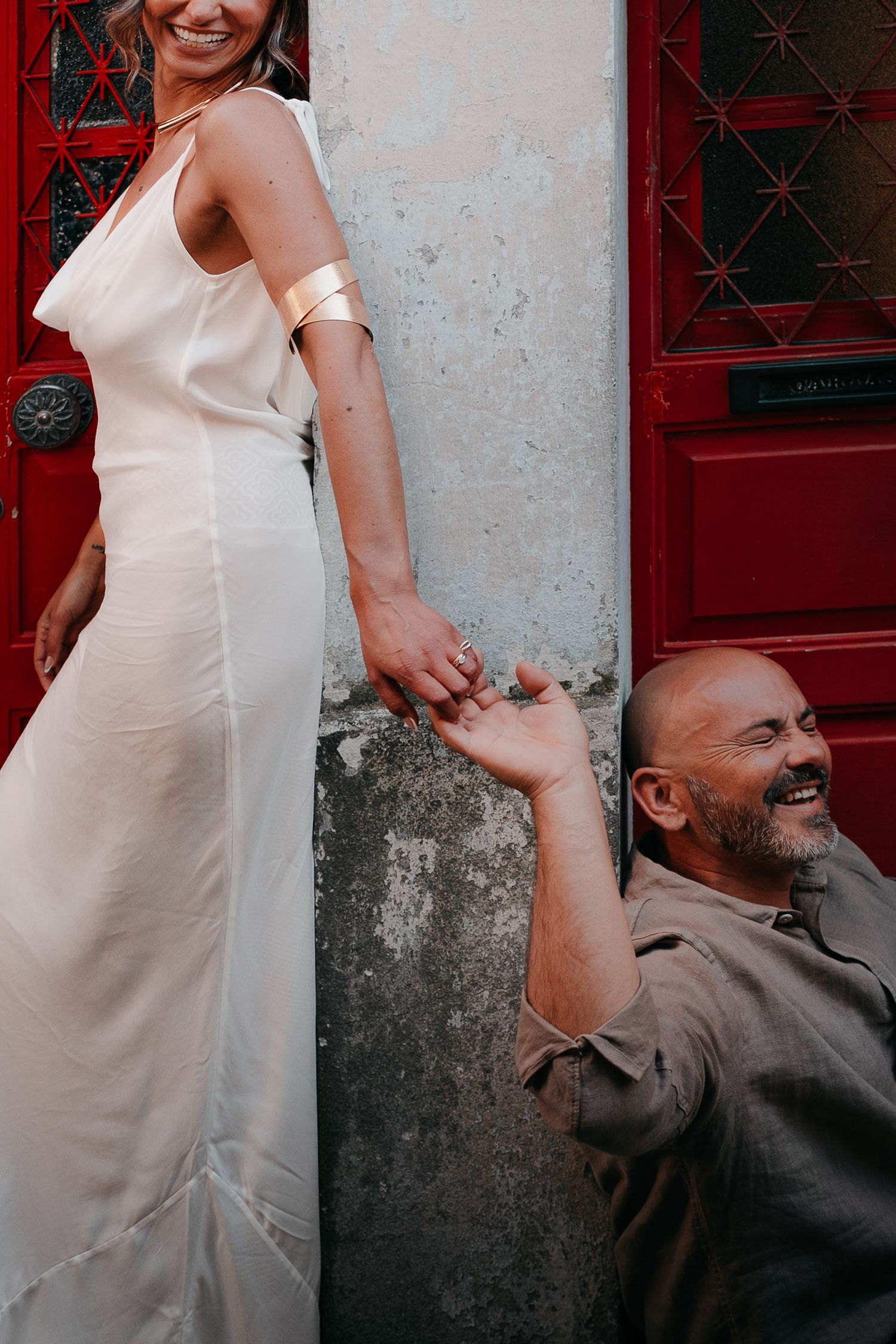 Bride in white gown holds hands with laughing groom seated by red door in romantic outdoor wedding photo.