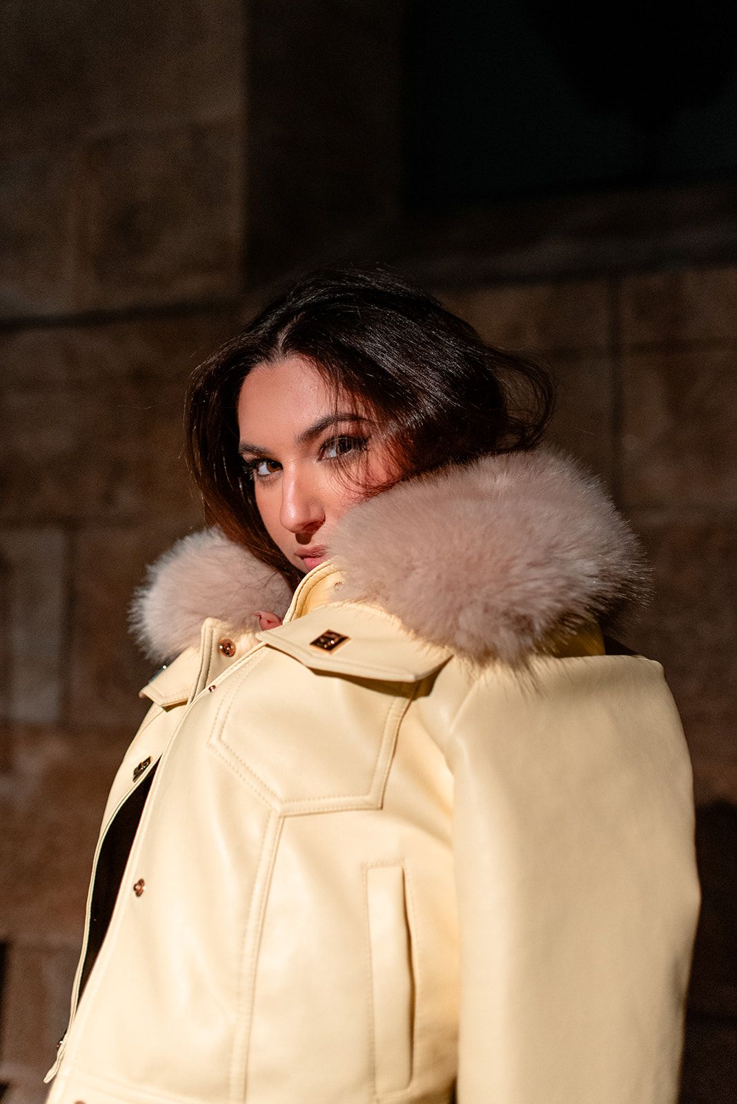 Fashion portrait featuring a cream colored fur-trimmed coat against stone walls.
