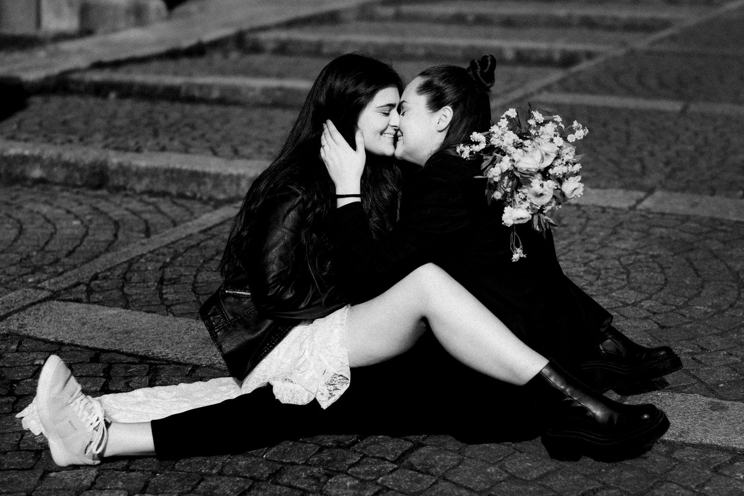 Black and white photo of two women sitting on cobblestones, sharing a kiss while holding a bouquet of flowers.