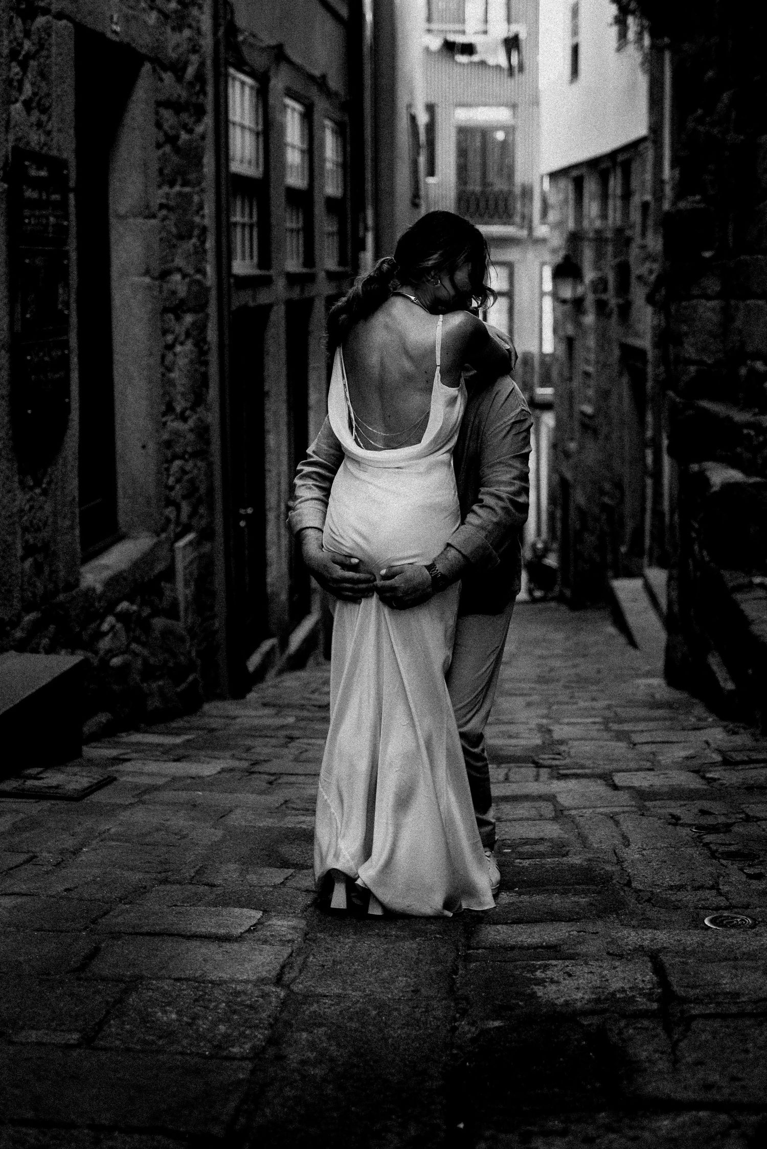 Black and white photo of couple embracing in a cobblestone alley, woman in backless gown.