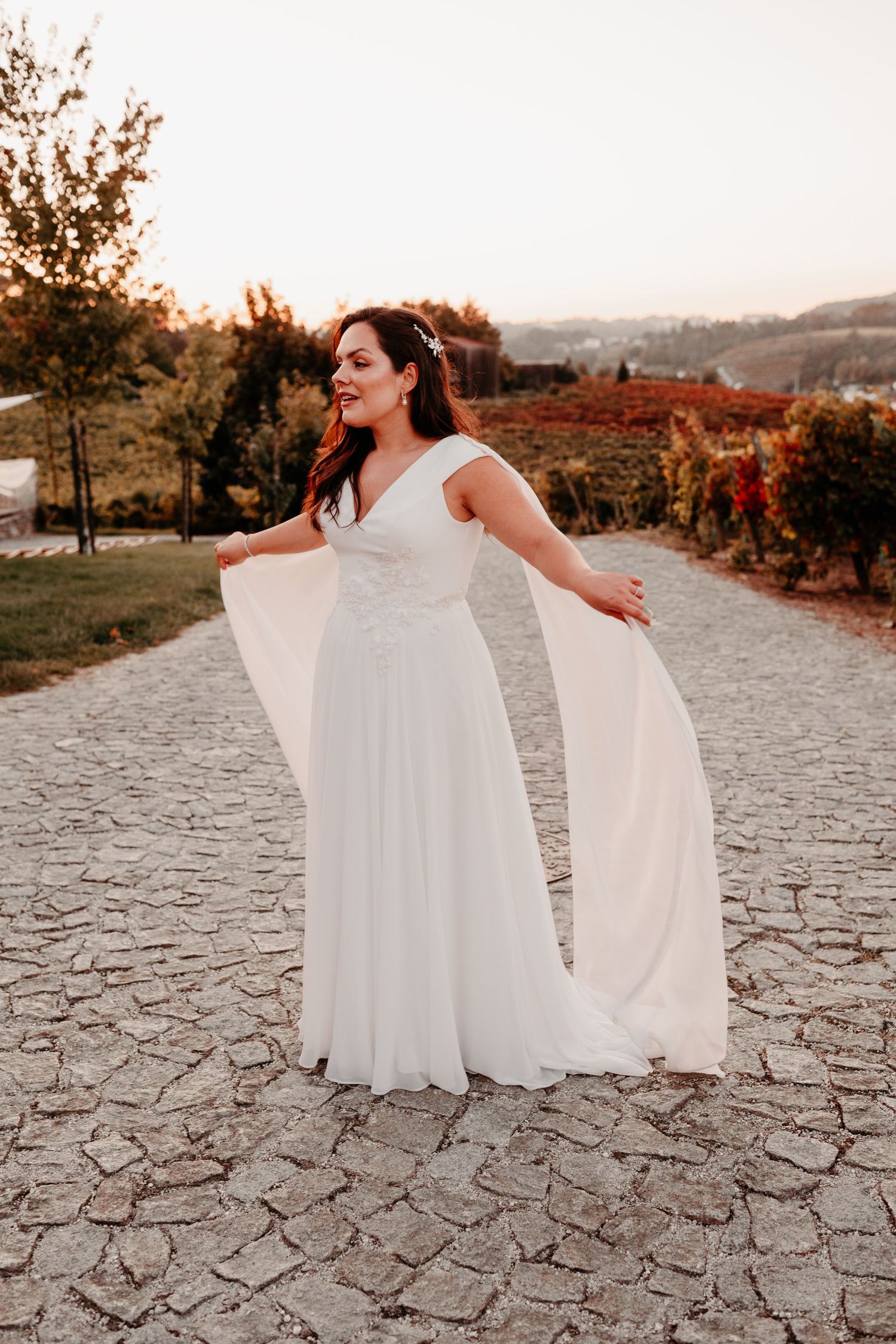 Bride in flowing white wedding gown with cape sleeves poses on cobblestone path surrounded by autumn vineyard at sunset.