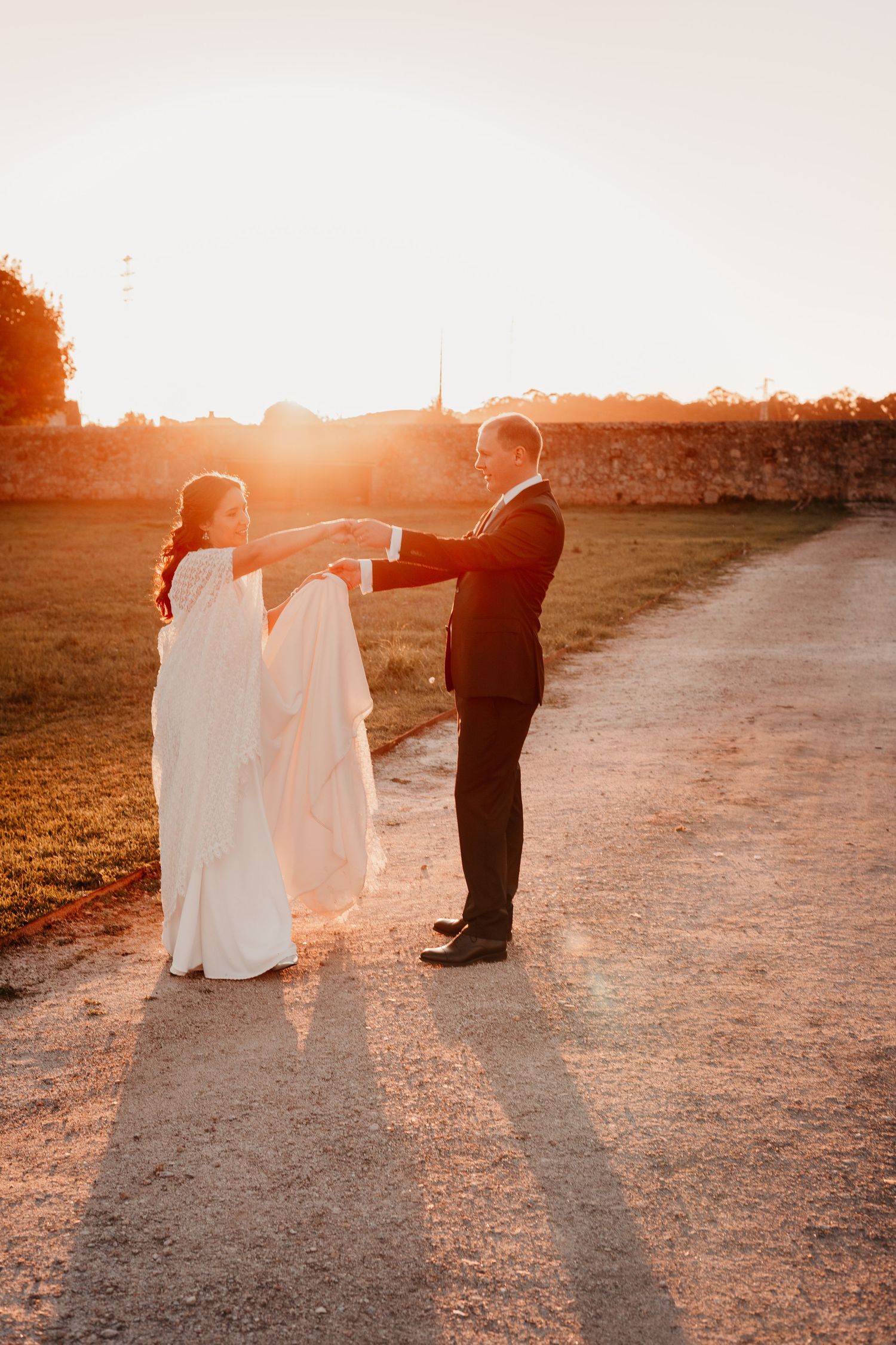 Bride and groom holding hands at golden sunset on a gravel path, bride in white gown, groom in black suit.