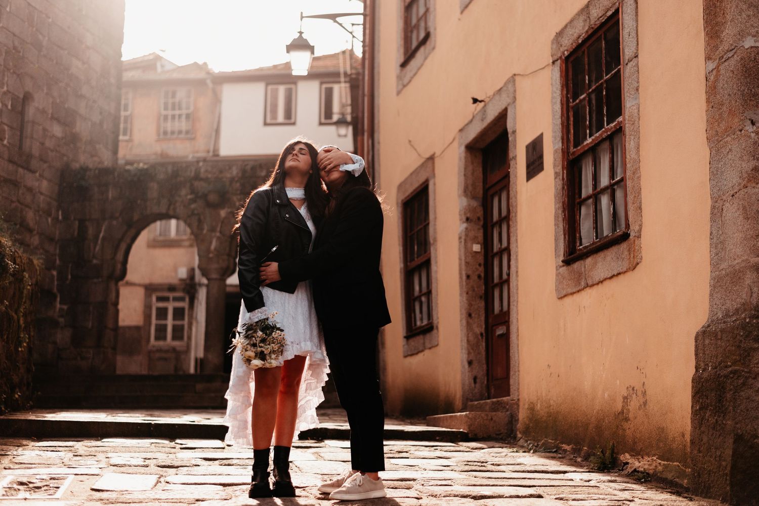 Couple in wedding attire embracing on cobblestone street in historic European town with warm sunlight.