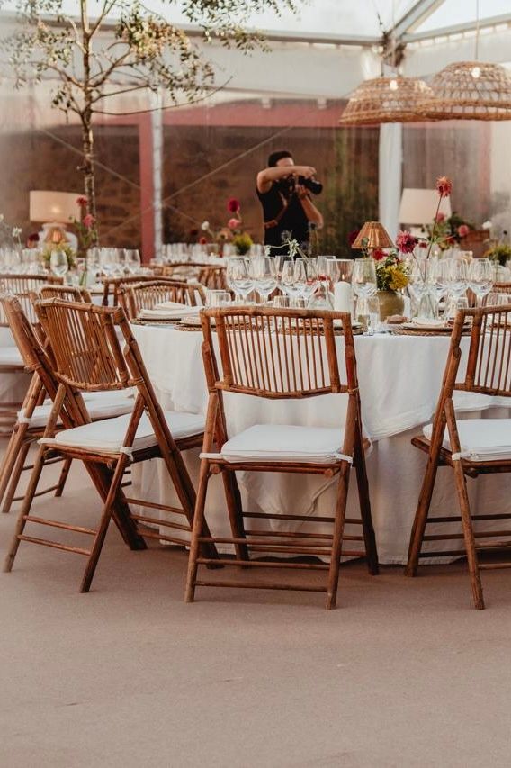 Elegant wedding reception with bamboo folding chairs and white cushions around round tables under a clear tent.
