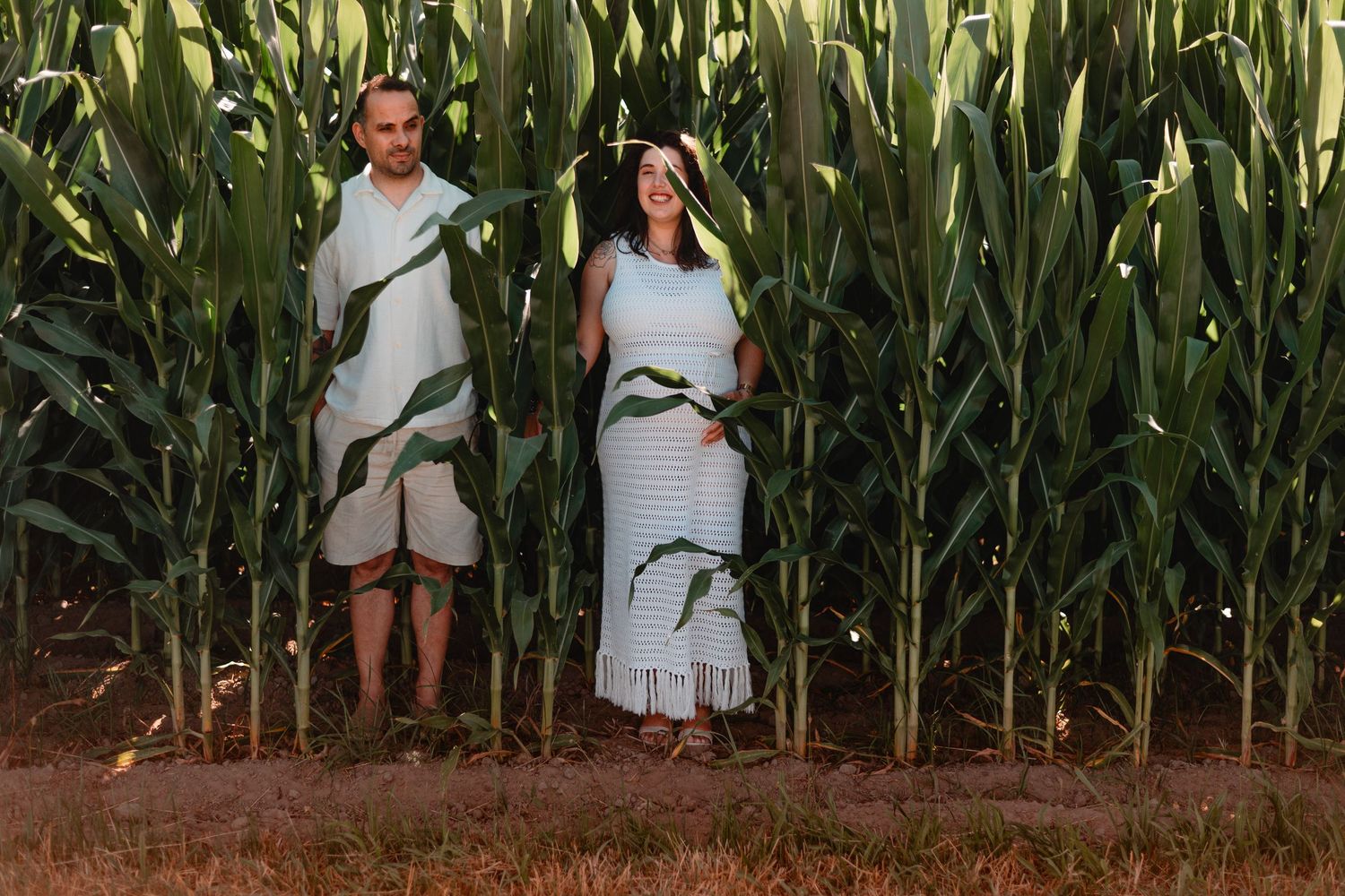 Couple poses among tall corn stalks in whimsical maternity photo shoot.