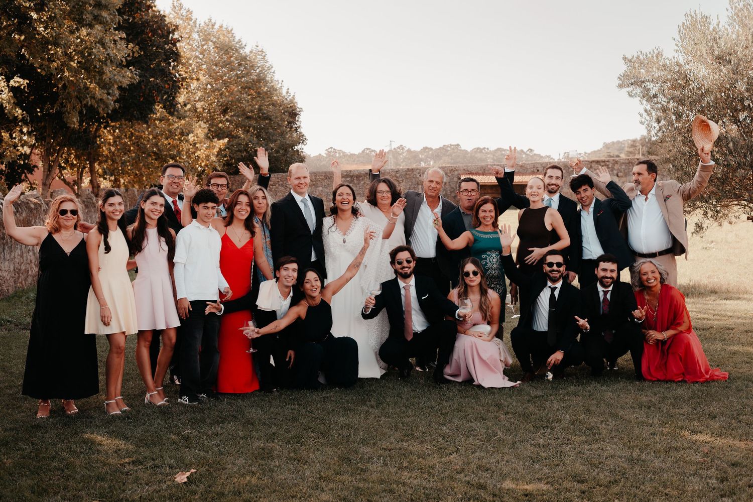 Large wedding group photo outdoors with bride and groom surrounded by smiling guests in formal attire near olive trees.