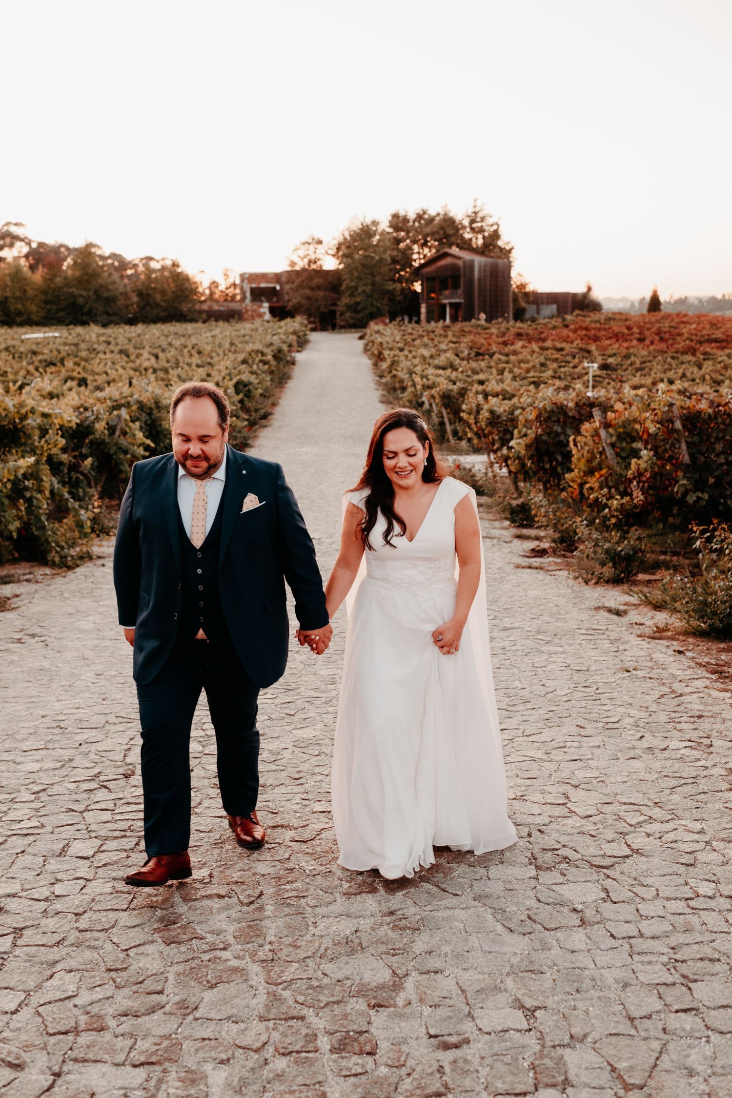 Bride and groom walk hand-in-hand along a cobblestone vineyard path at sunset, wearing white gown and navy suit.