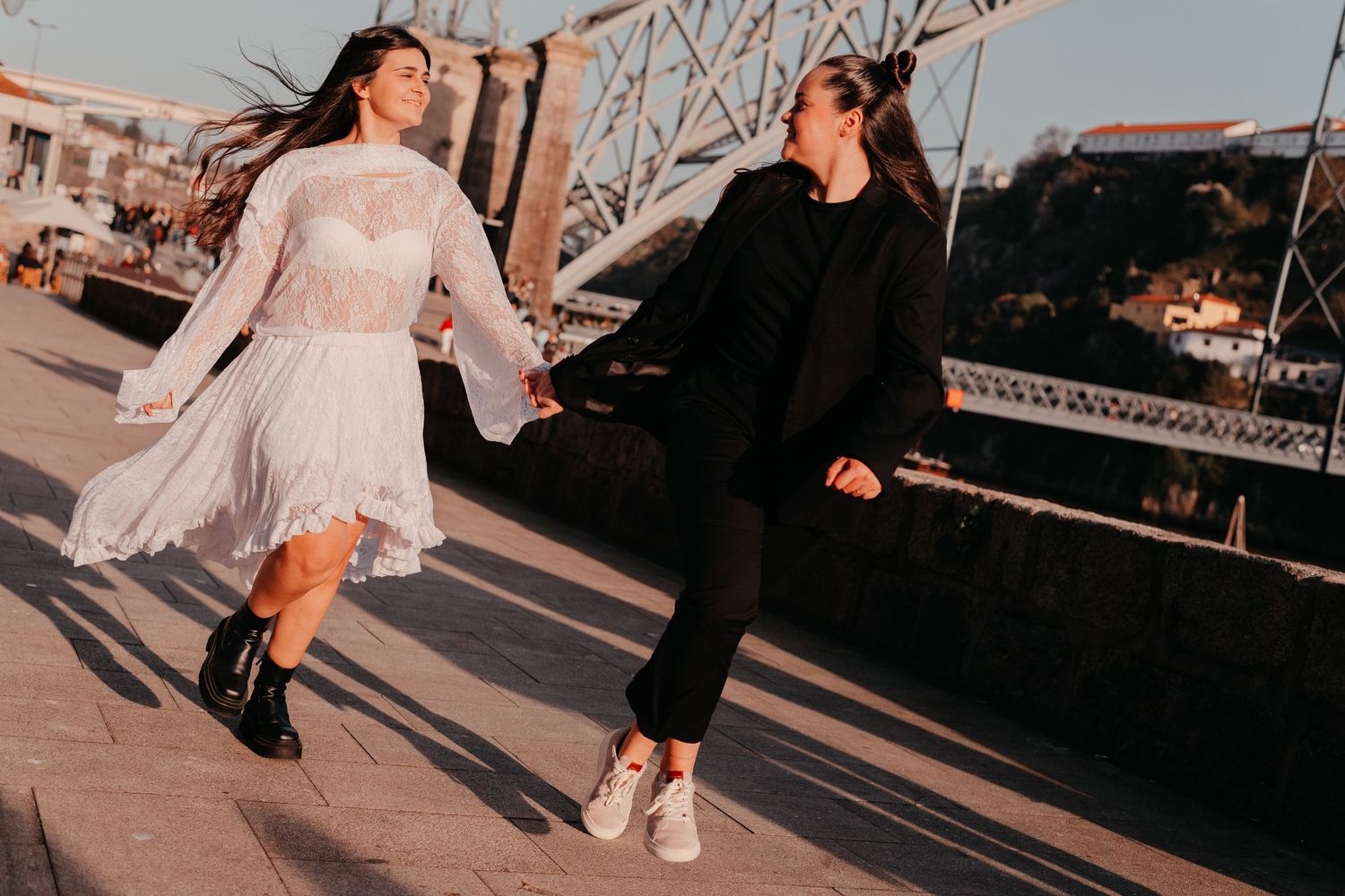 Two women holding hands and laughing while walking on a bridge at sunset, one in white dress and one in black outfit.
