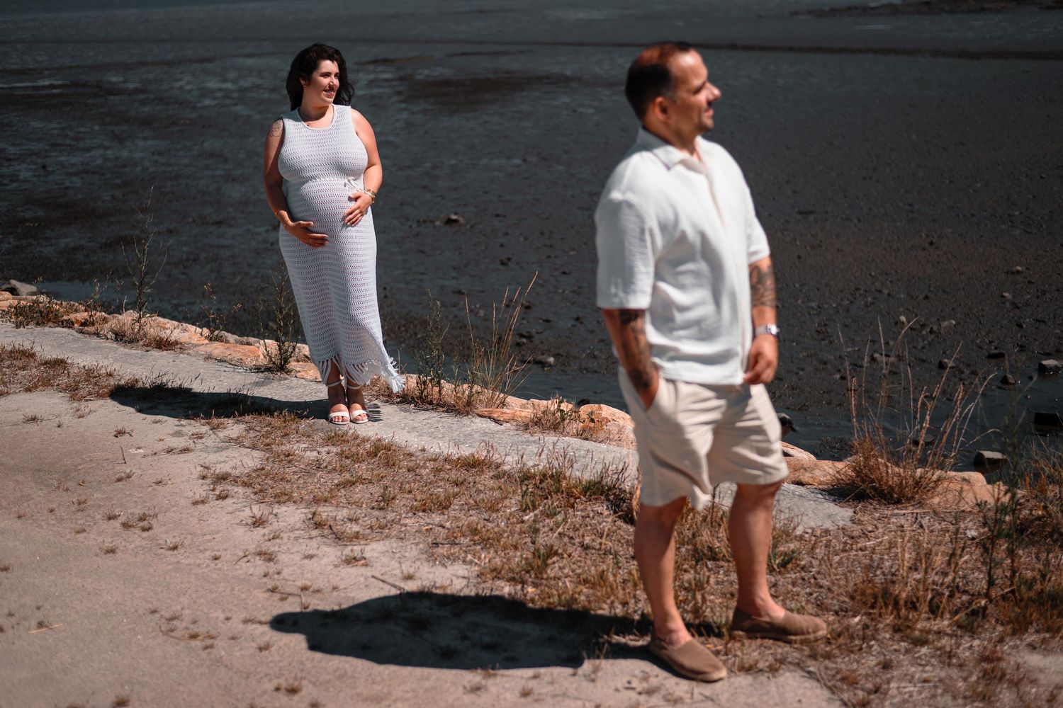 A couple in white outfits stands on a sandy beach path against a dark water backdrop.