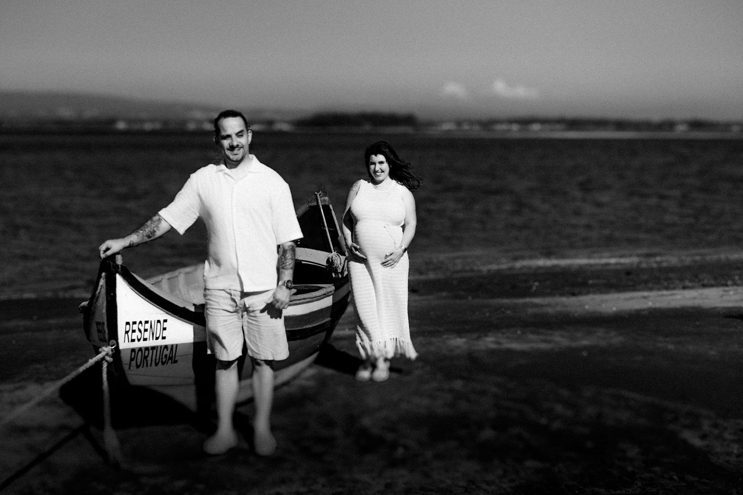 Black and white beach portrait with couple standing beside vintage wooden boat.