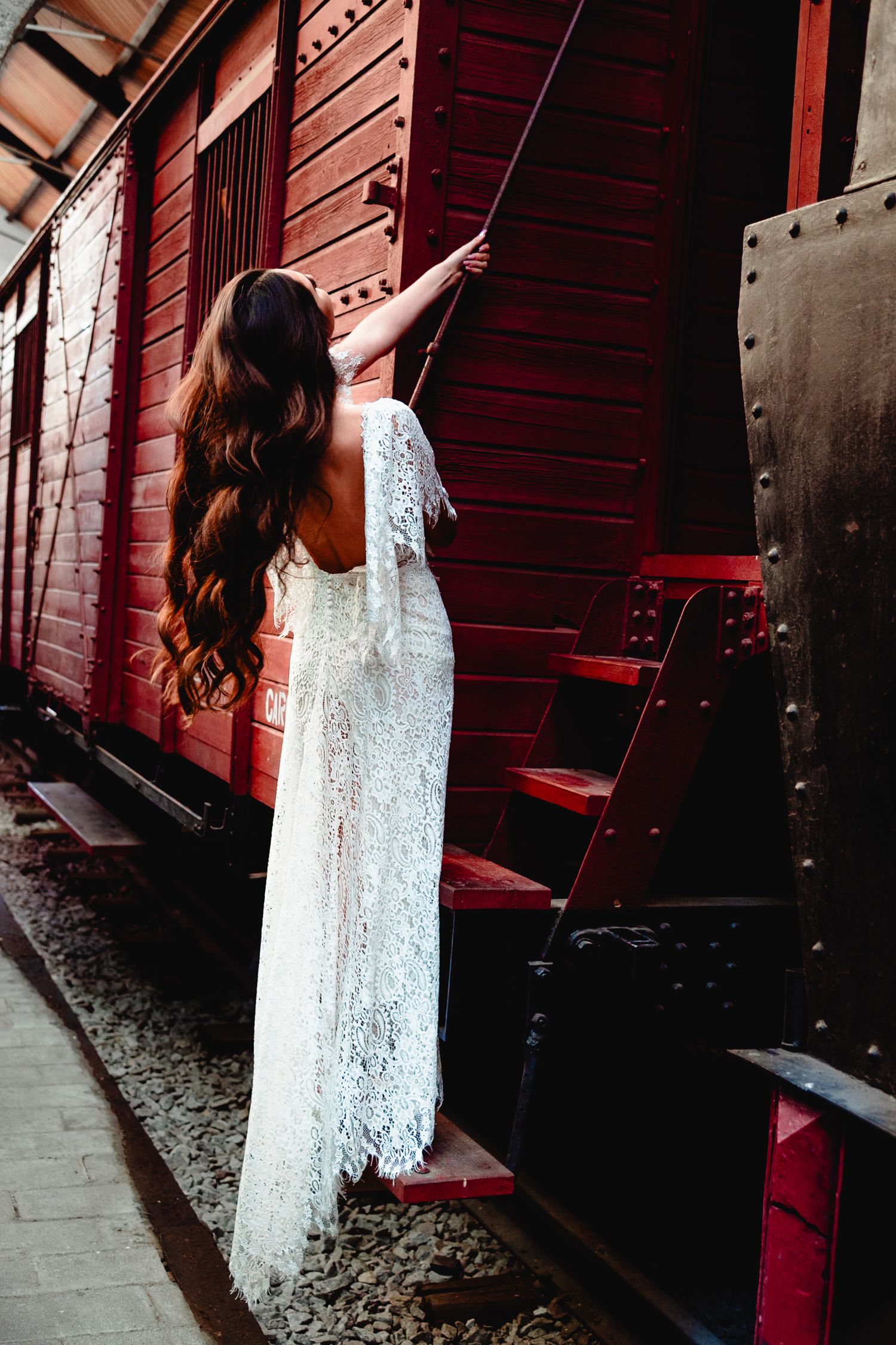 Elegant bridal photo with long white lace dress against rustic red train car exterior.