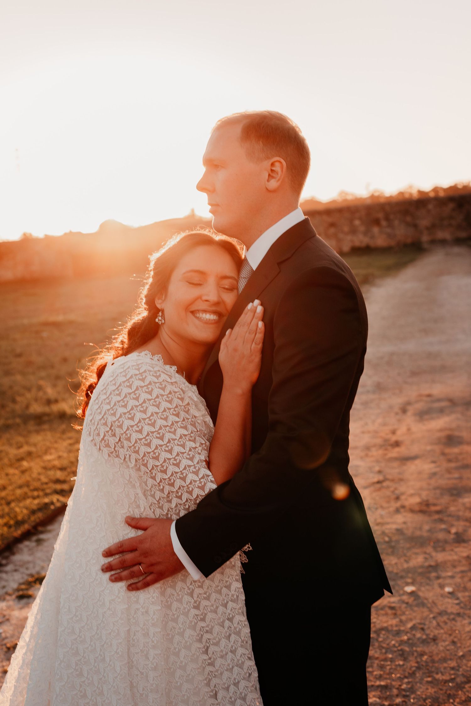 Bride and groom embrace at golden hour sunset, bride in lace dress smiling joyfully during romantic outdoor wedding portrait.