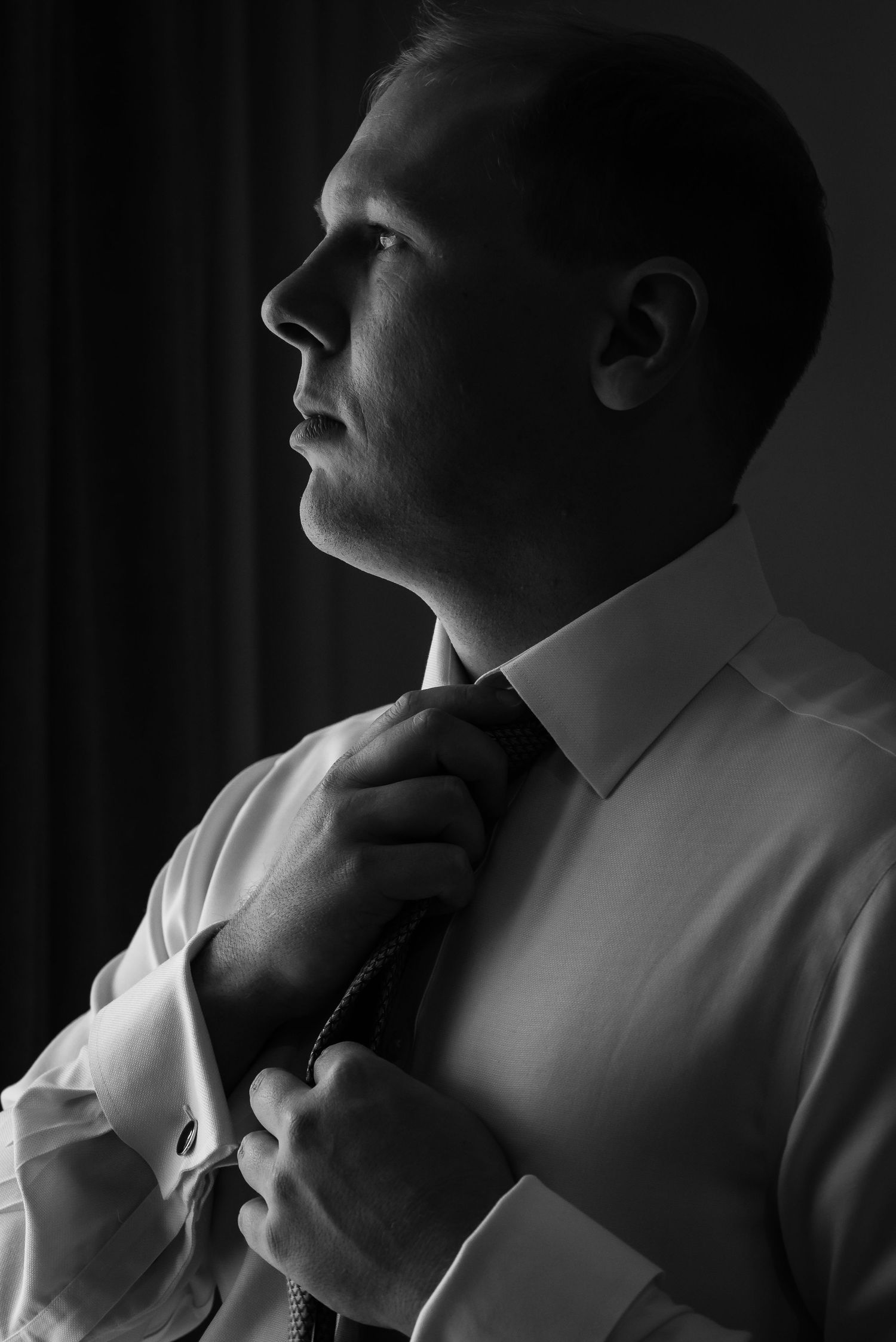 Black and white portrait of a man in a white dress shirt adjusting his tie, dramatic side lighting.