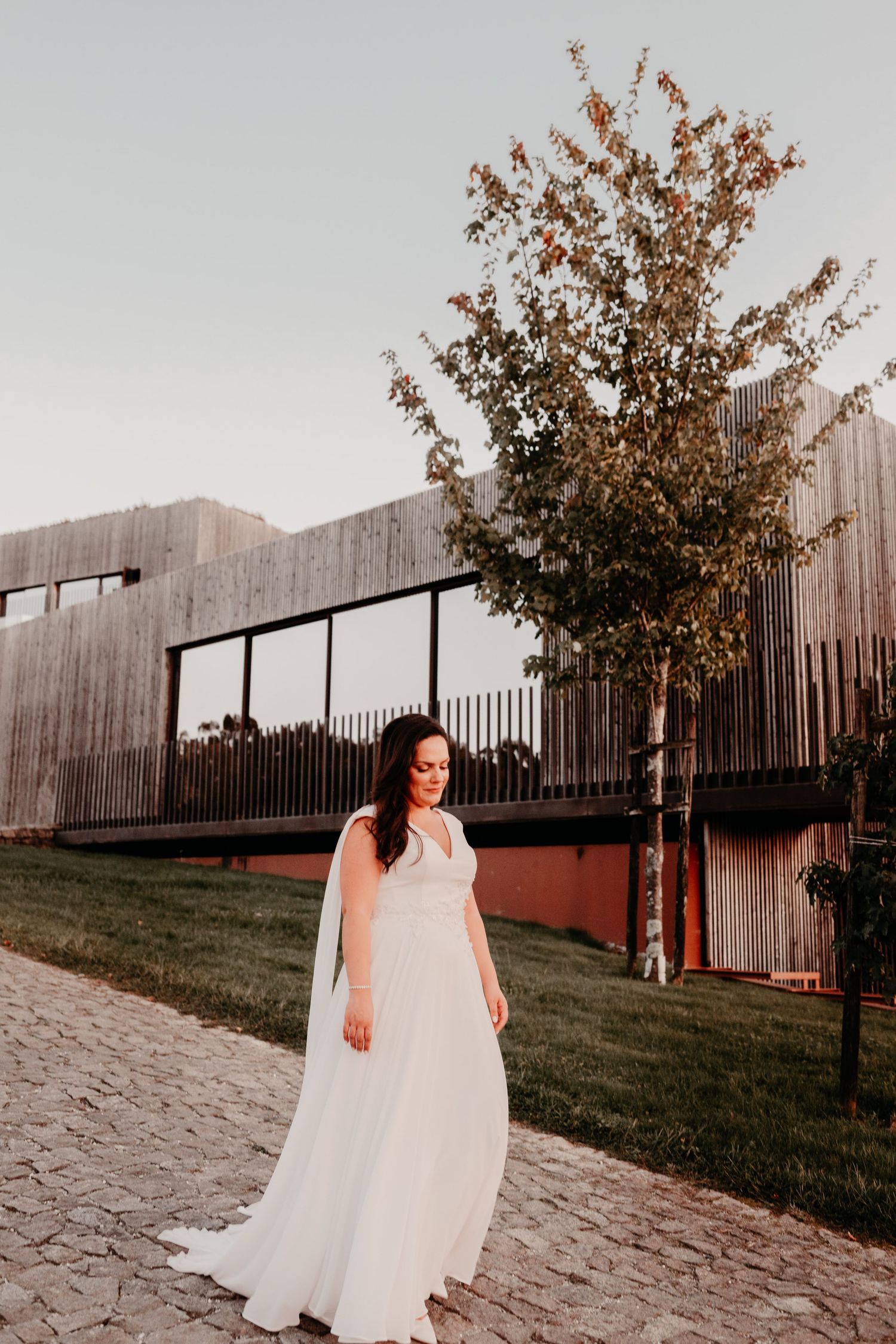 Bride in white gown with veil stands on cobblestone path before modern wooden building and tree at golden hour.