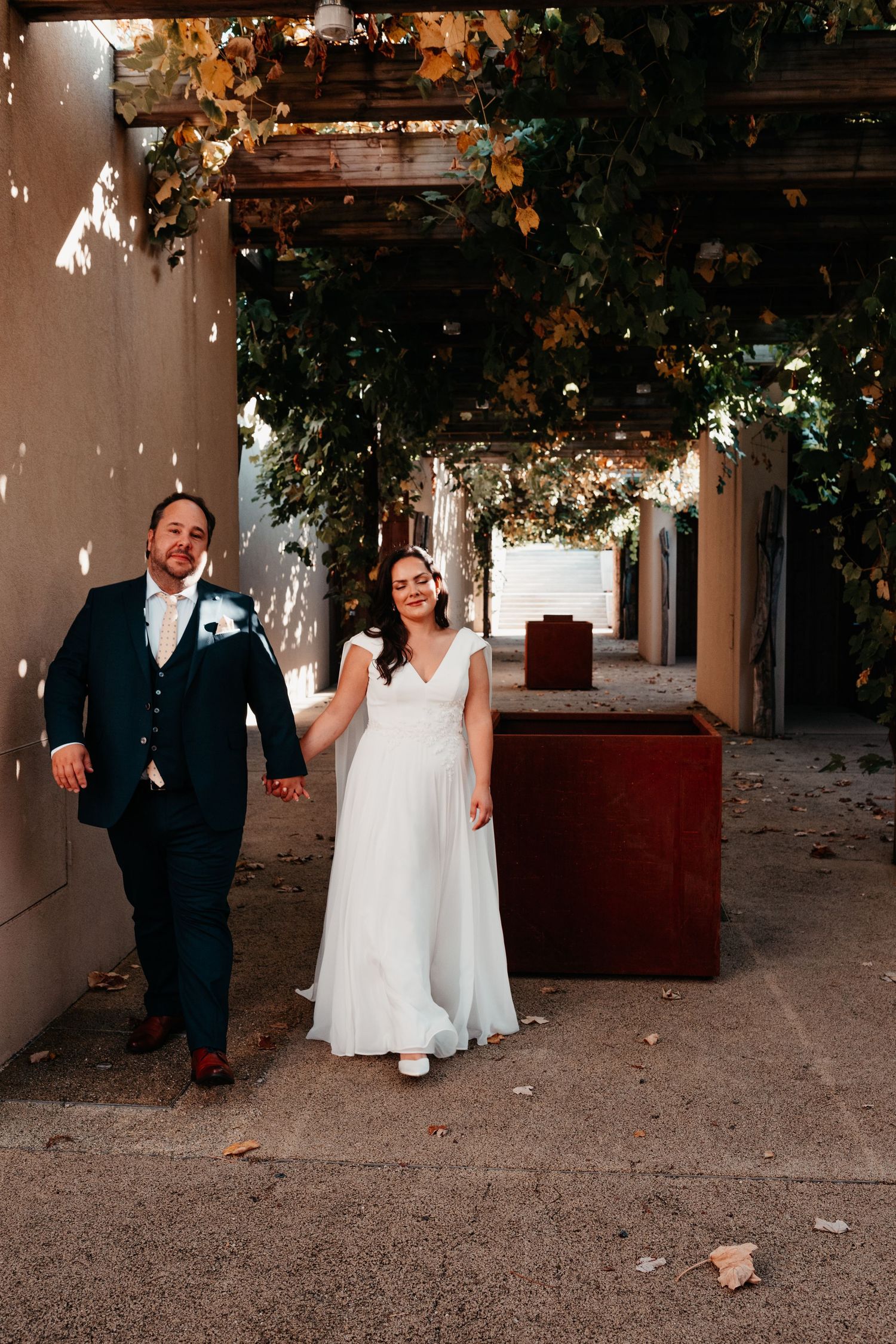 A bride and groom walk hand in hand under a vine-covered pergola in autumn, smiling joyfully on their wedding day.