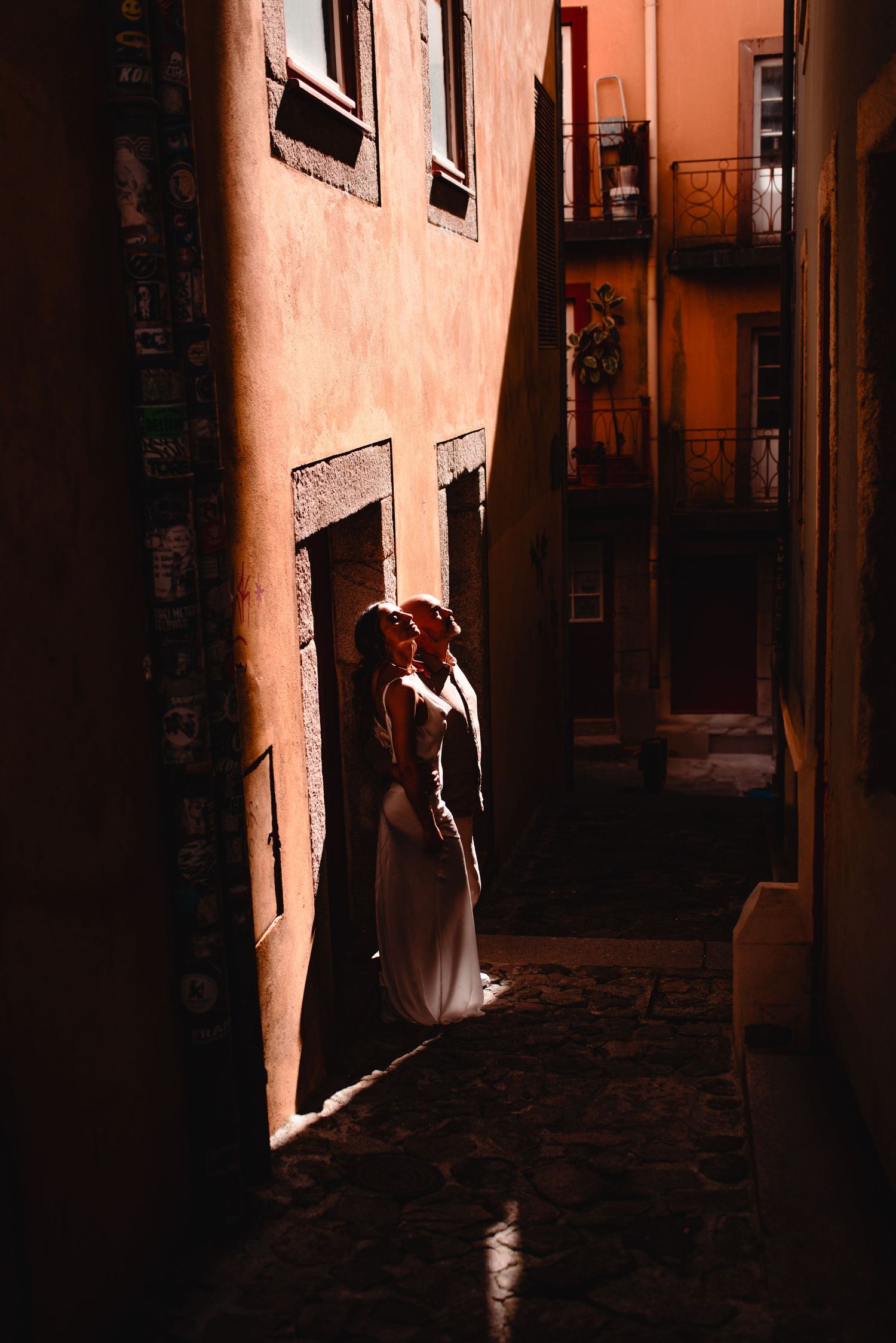 A figure in a white dress stands in dramatic lighting against a terracotta wall in a narrow European alley.