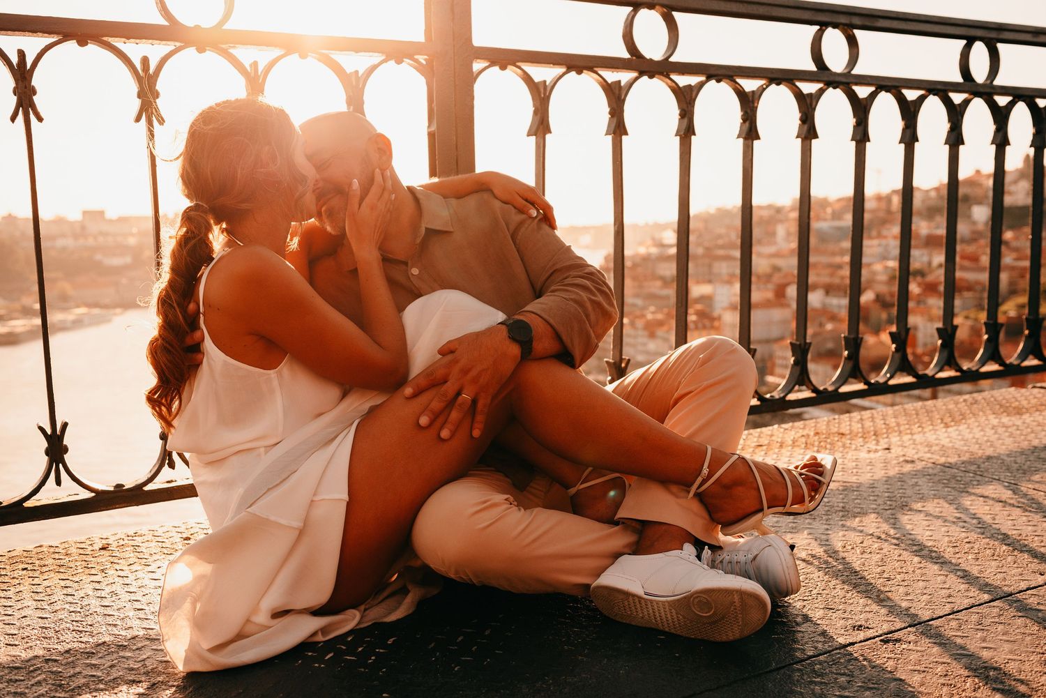 Romantic couple sitting together on a balcony at golden sunset, sharing a tender kiss outdoors.