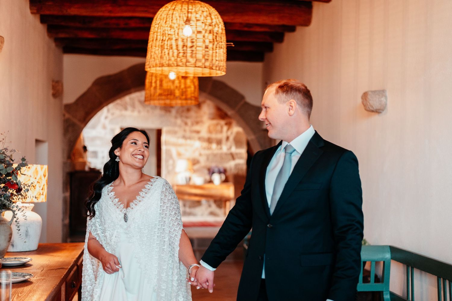 A bride and groom hold hands and smile at each other inside a rustic venue with stone arch and woven pendant light.