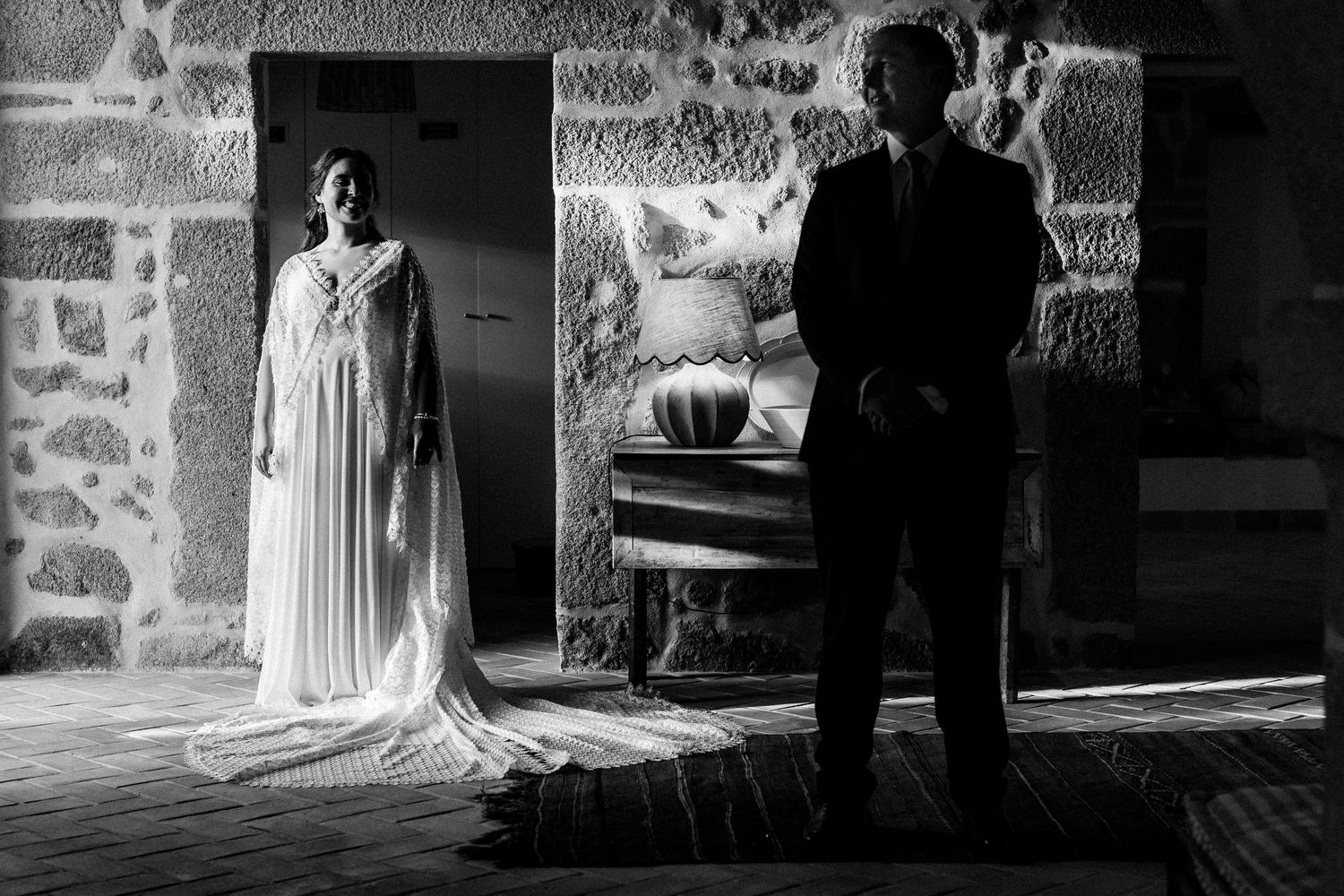 Black and white wedding photo of bride in flowing gown and groom in suit standing apart in dramatic stone setting.