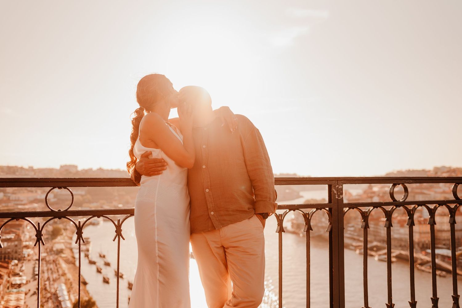 Romantic couple embracing on a sunlit balcony overlooking a city river at golden hour.