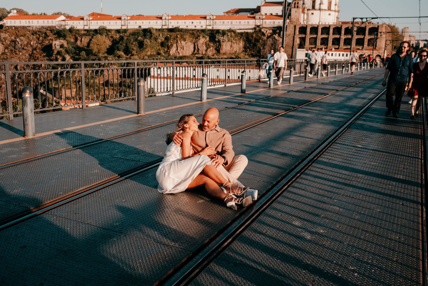 Couple sitting together on a sunlit bridge with tram tracks in Porto, Portugal, sharing a romantic moment.