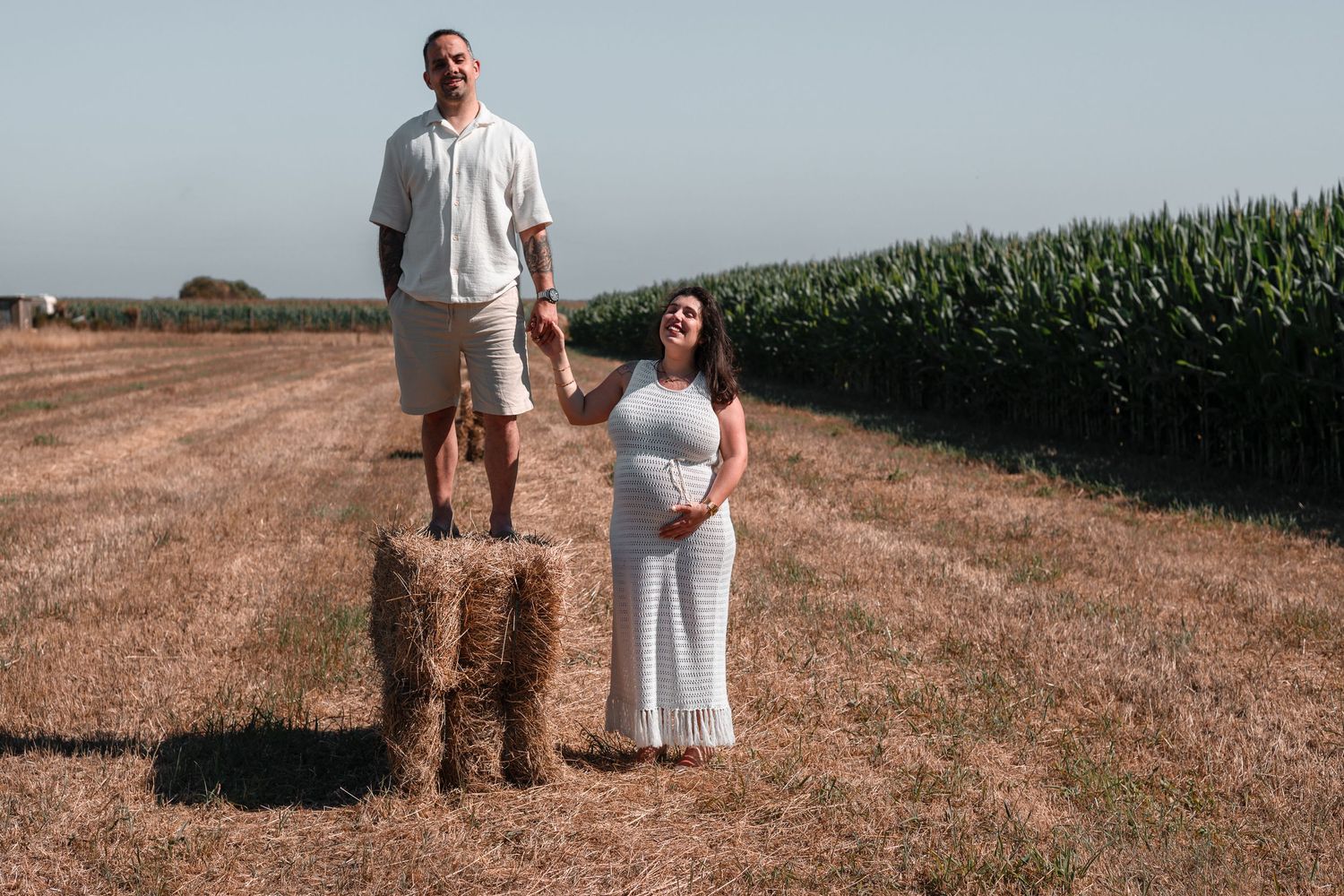 A couple poses in a sunlit field with tall corn stalks in the background.