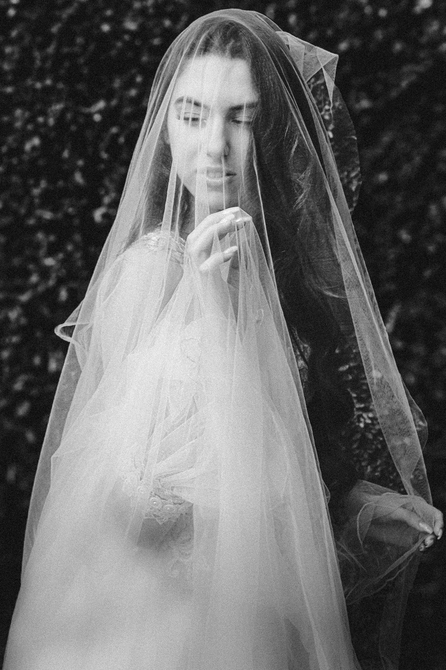 Ethereal black and white portrait of a bride covered in delicate sheer veil against a dark background.