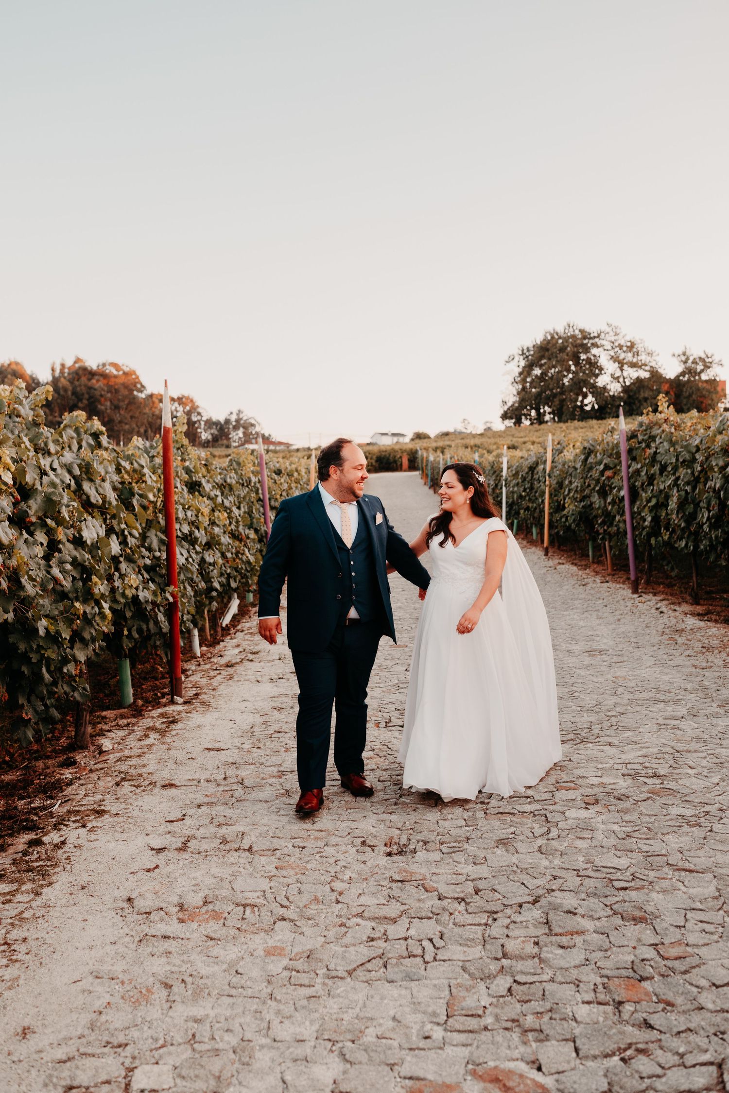 Bride and groom walk hand in hand through a sunlit vineyard path, smiling at each other on their wedding day.