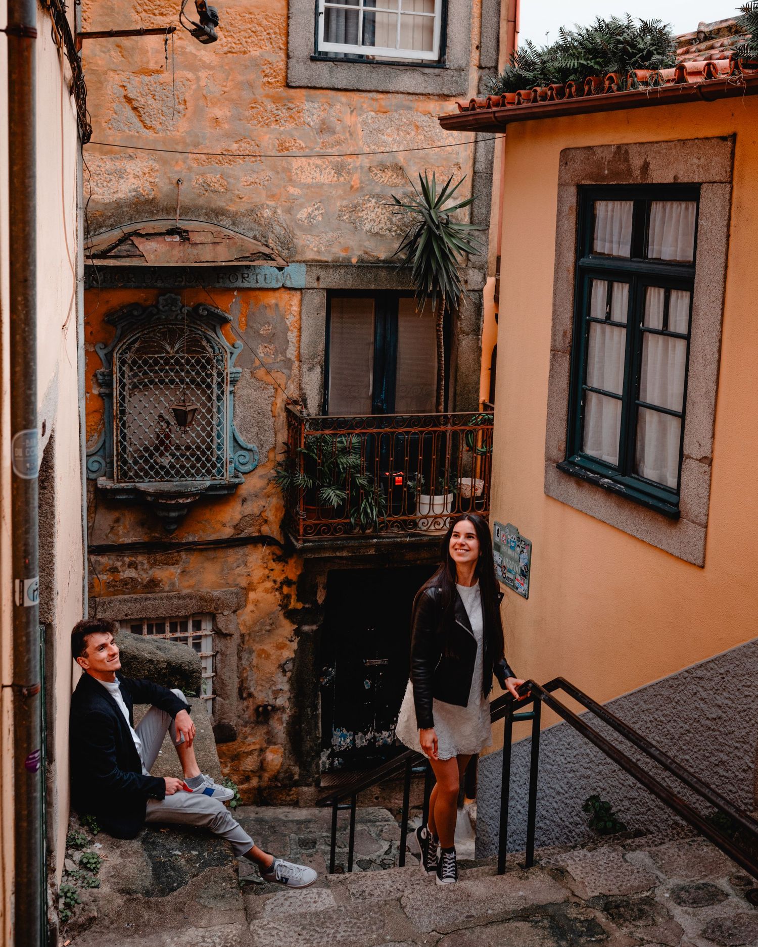 Couple posing on old stone steps in Porto, Portugal, with rustic colorful buildings and lush greenery in background.