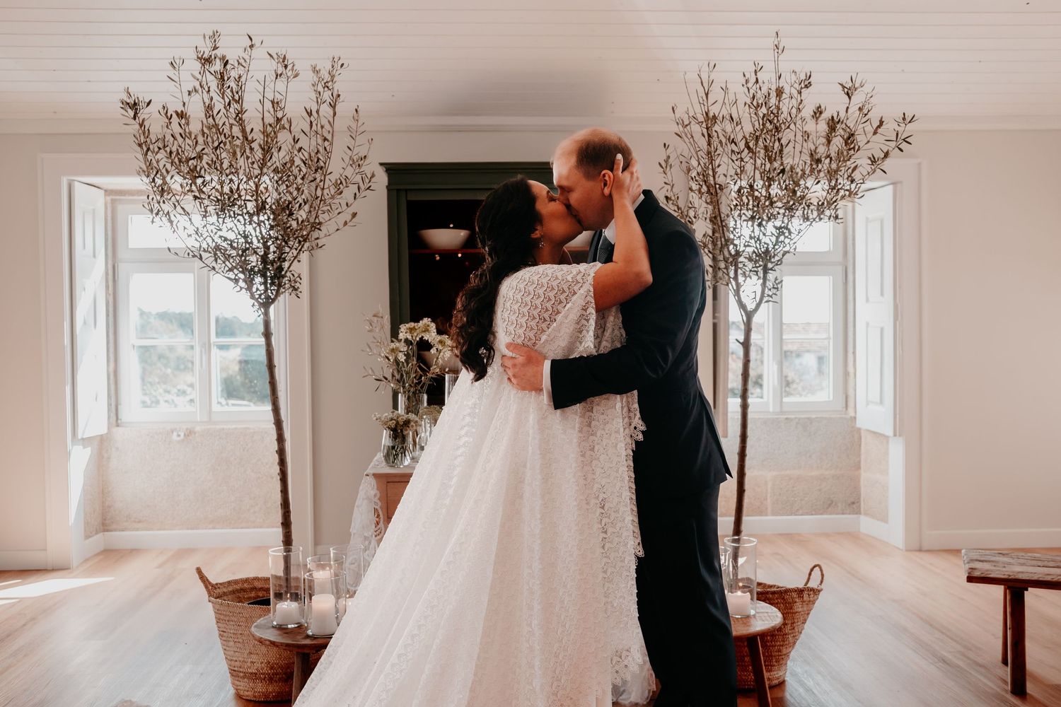 Bride and groom share first kiss at elegant indoor wedding ceremony with olive trees and white walls.