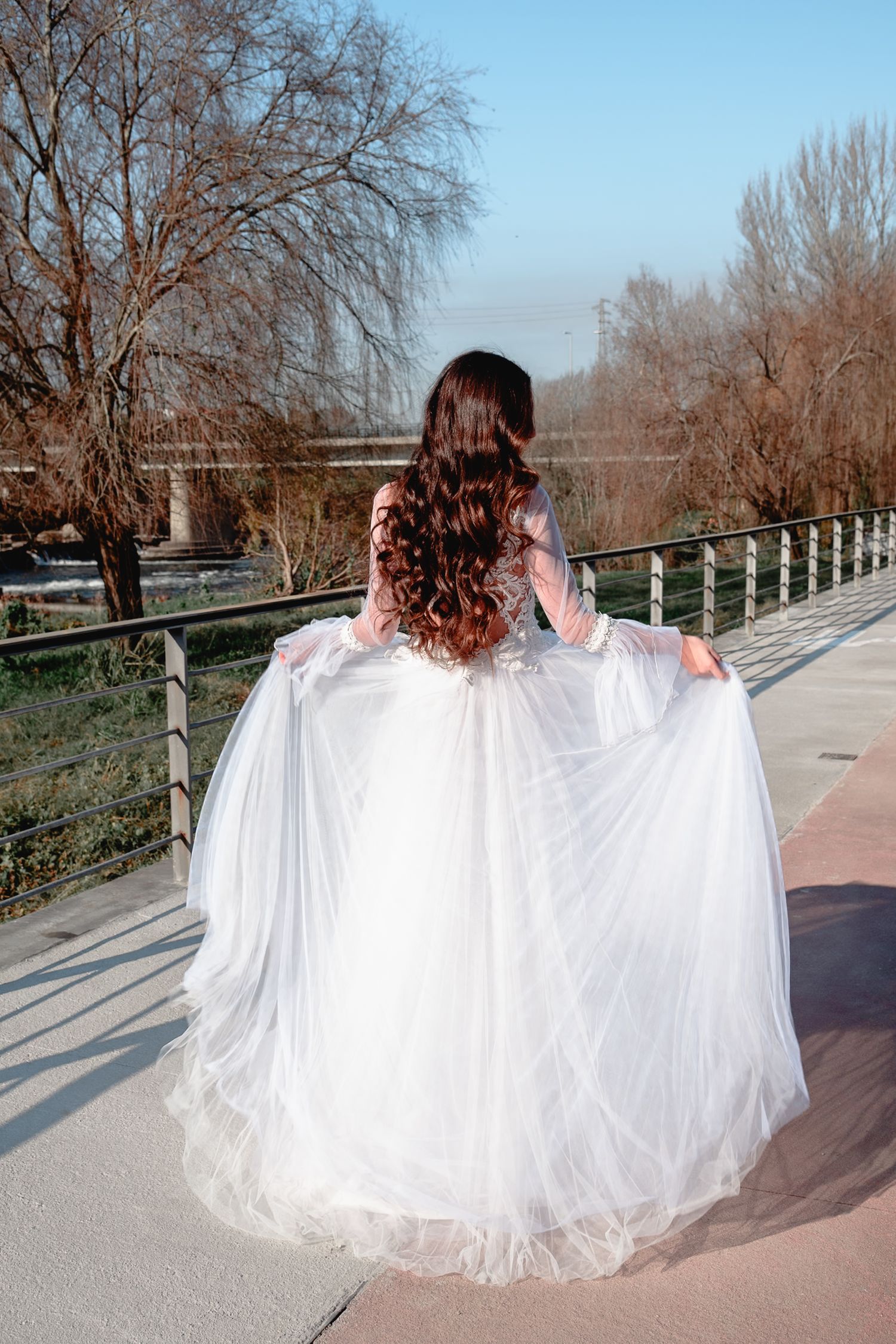 A billowing white wedding gown photographed along a riverside path with bare trees.