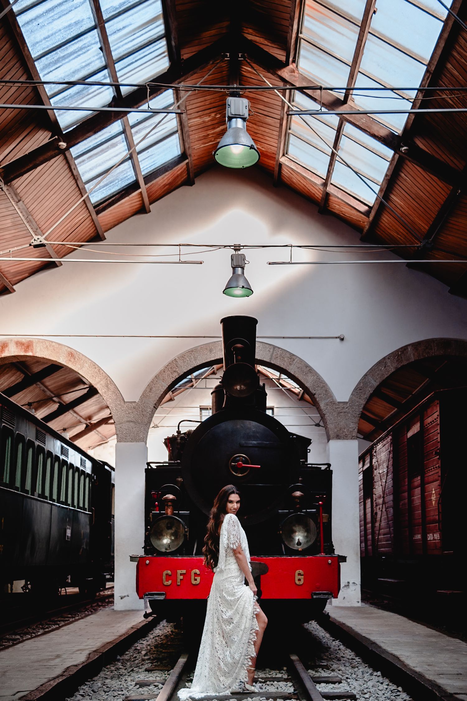 Bride in white dress standing in front of vintage steam locomotive under glass ceiling station.